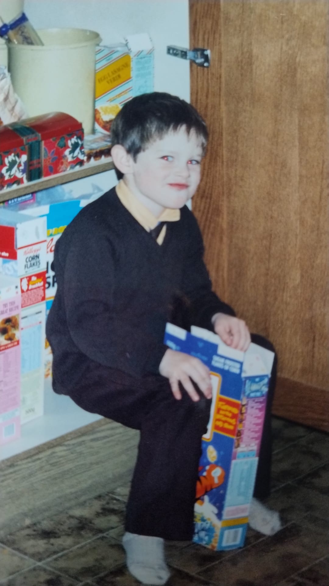 James Hacking as a young boy in school uniform, crouching in the family kitchen beside cereal boxes in the mid 1990s