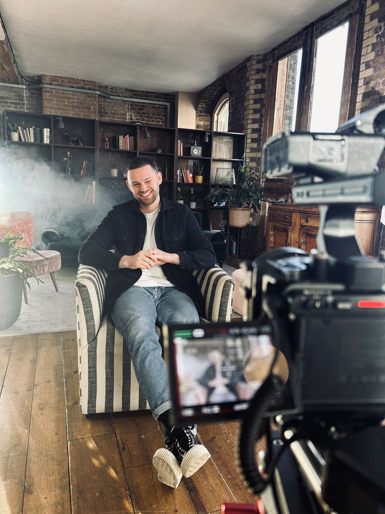 James Hacking sitting in an armchair, smiling while being filmed on camera in a studio